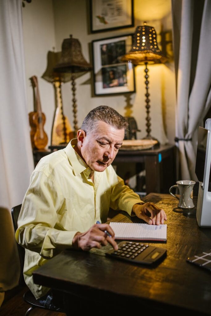 Elderly man working on calculations and taking notes in home study with warm lighting.